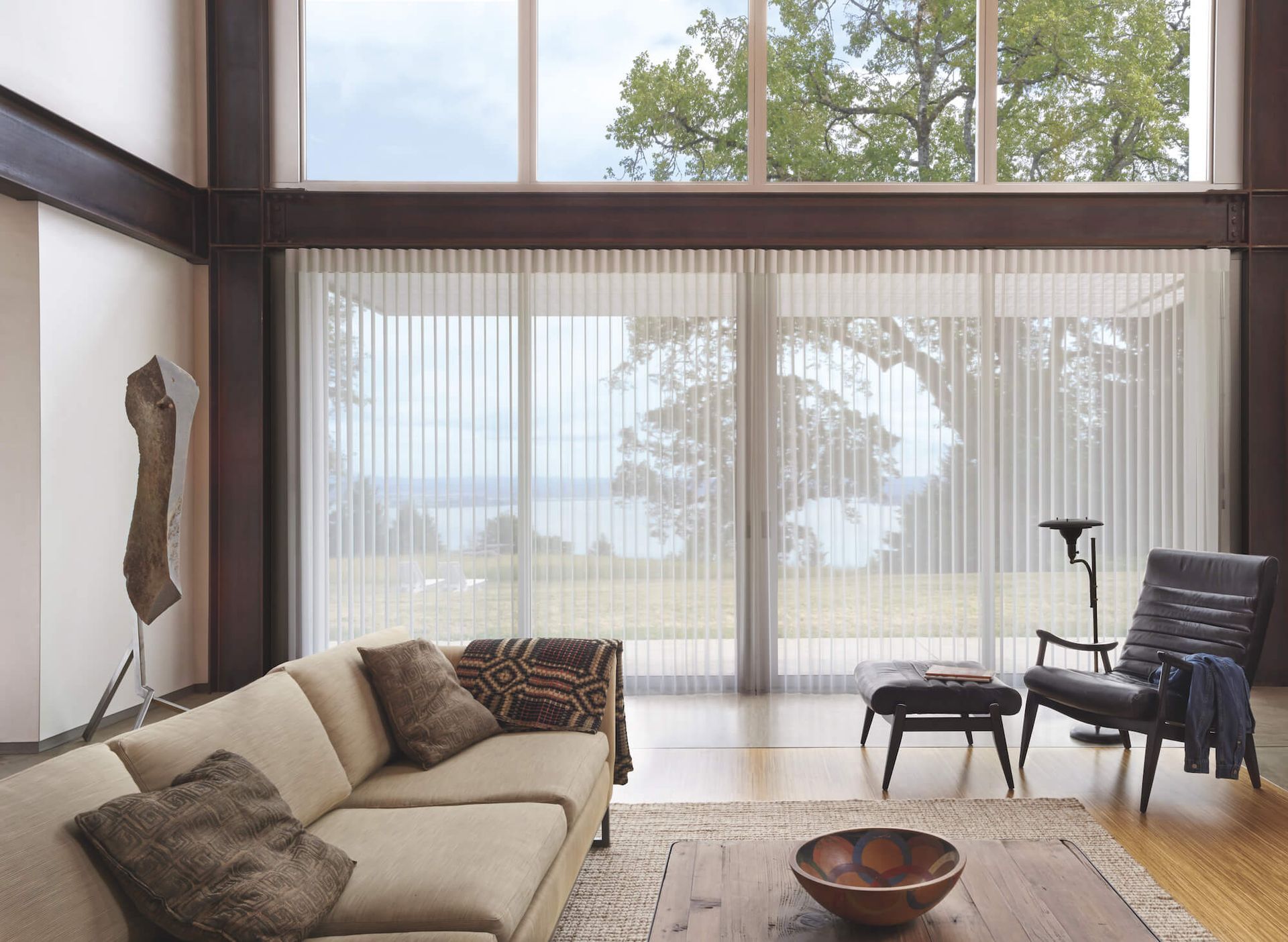 Modern living room with beige sofa, black chairs, wood floor, and large windows with sheer curtains.