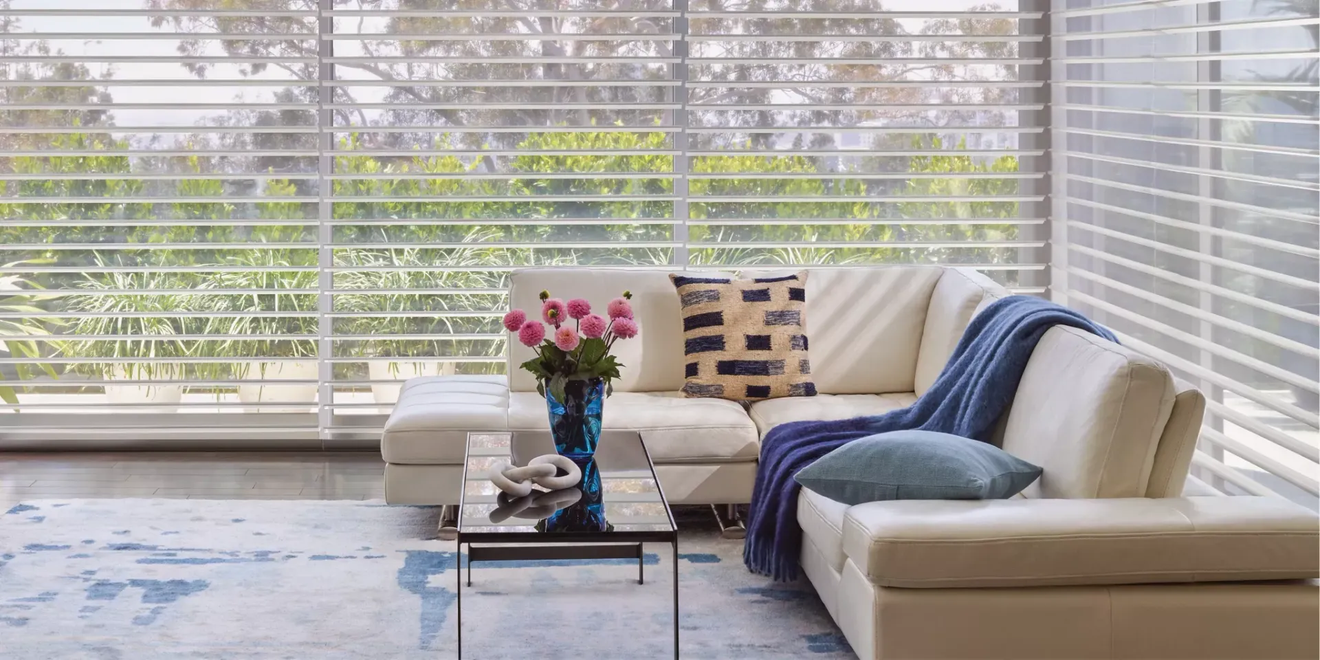 Bright living room with white sofas, blue throw, coffee table, and large windows with blinds.