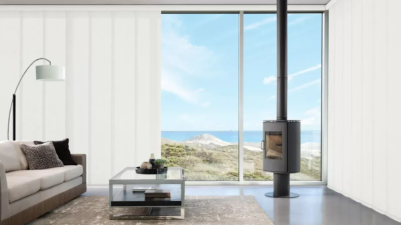 Modern living room with sofa, coffee table, floor lamp, wood stove, and large windows overlooking the sea