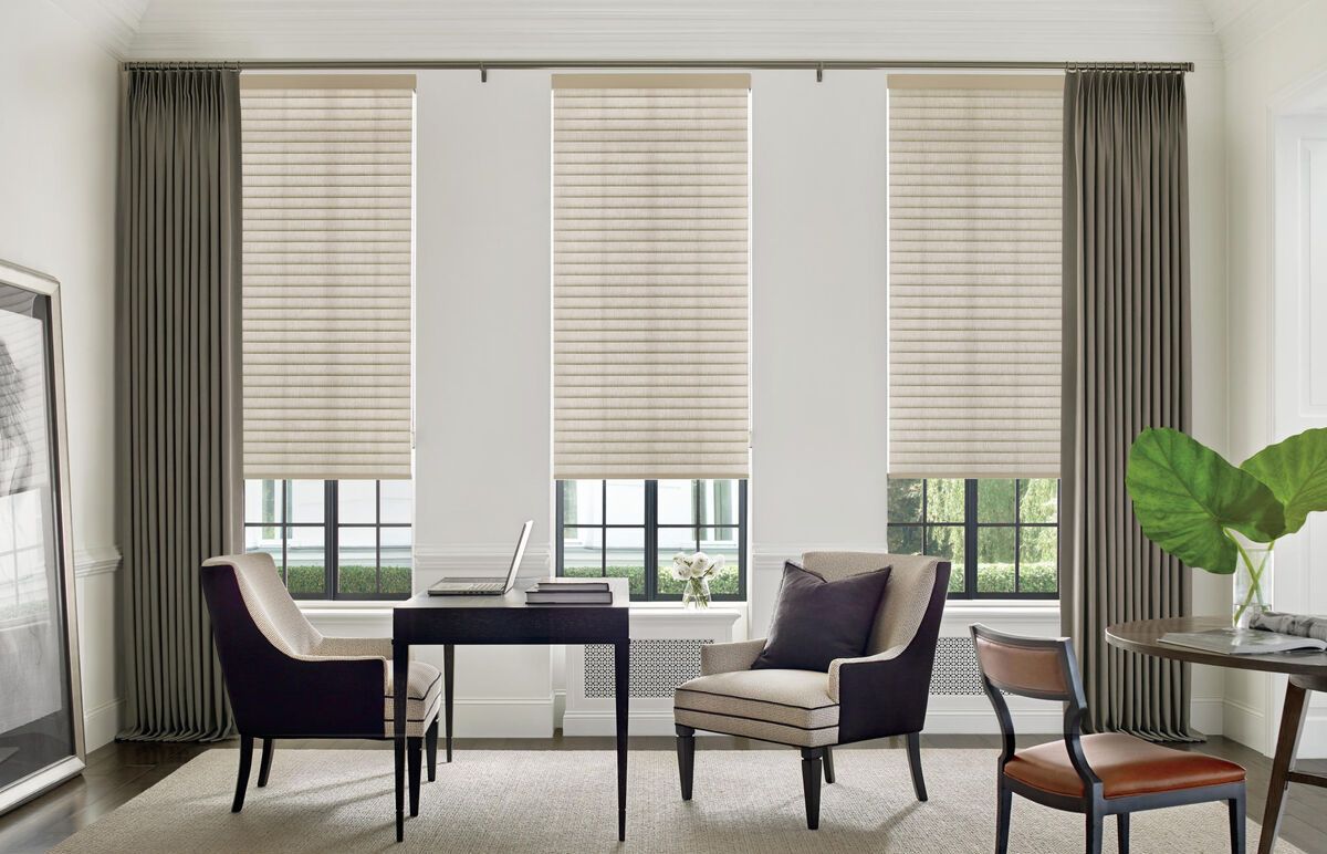 Elegant living room with neutral curtains, two chairs, a desk, and large windows with woven shades.