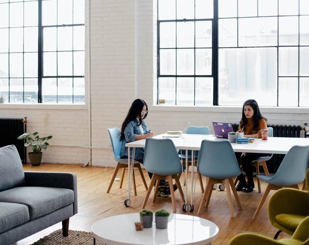 Two women are sitting at a table with laptops in a living room.