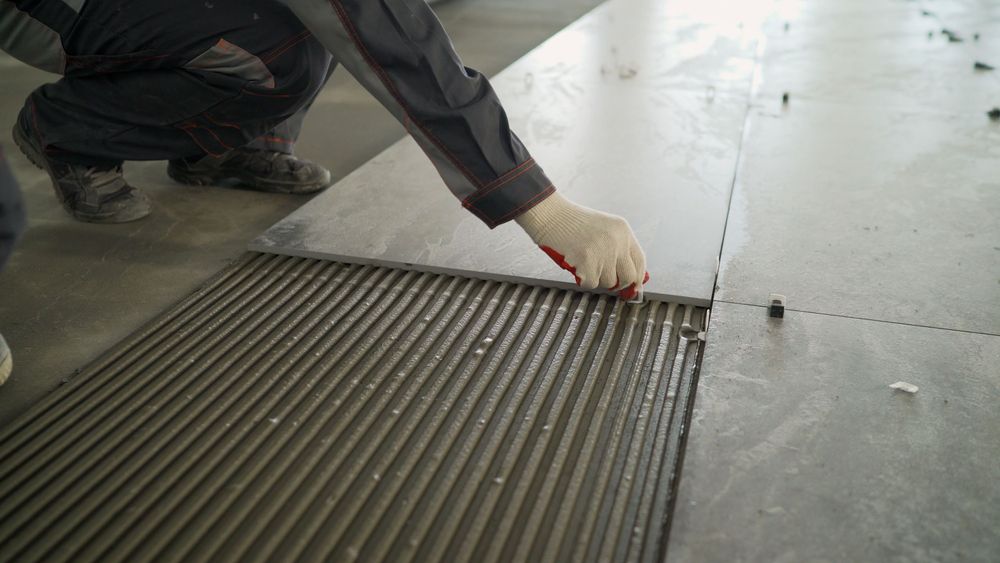 Person kneeling, placing a tile over adhesive; gray and neutral tones dominate.