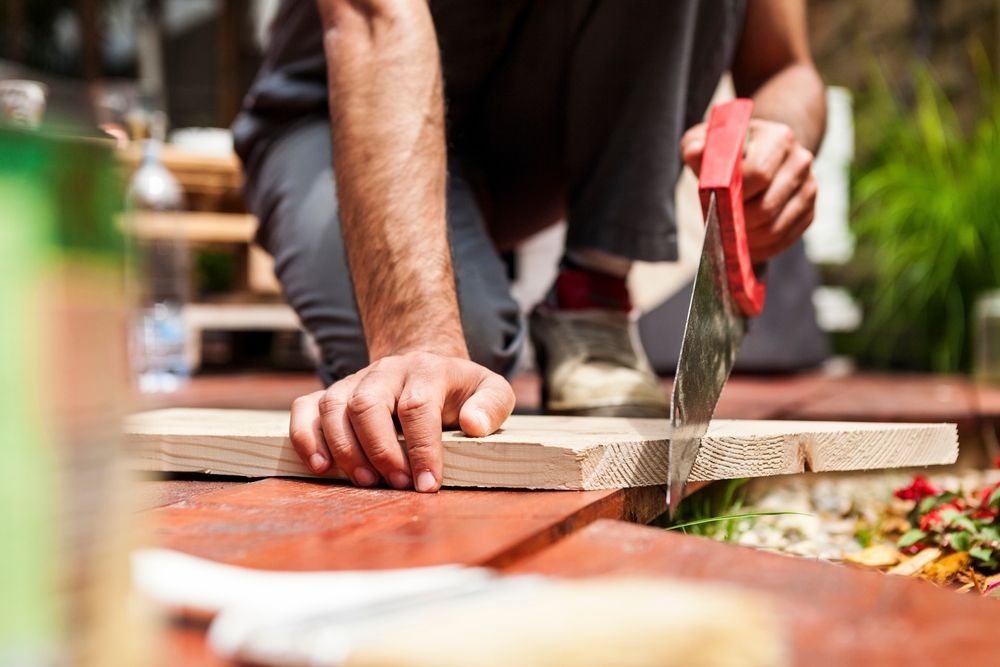 Person kneeling, sawing wood on a wooden surface outdoors, red handle saw, sunny day.