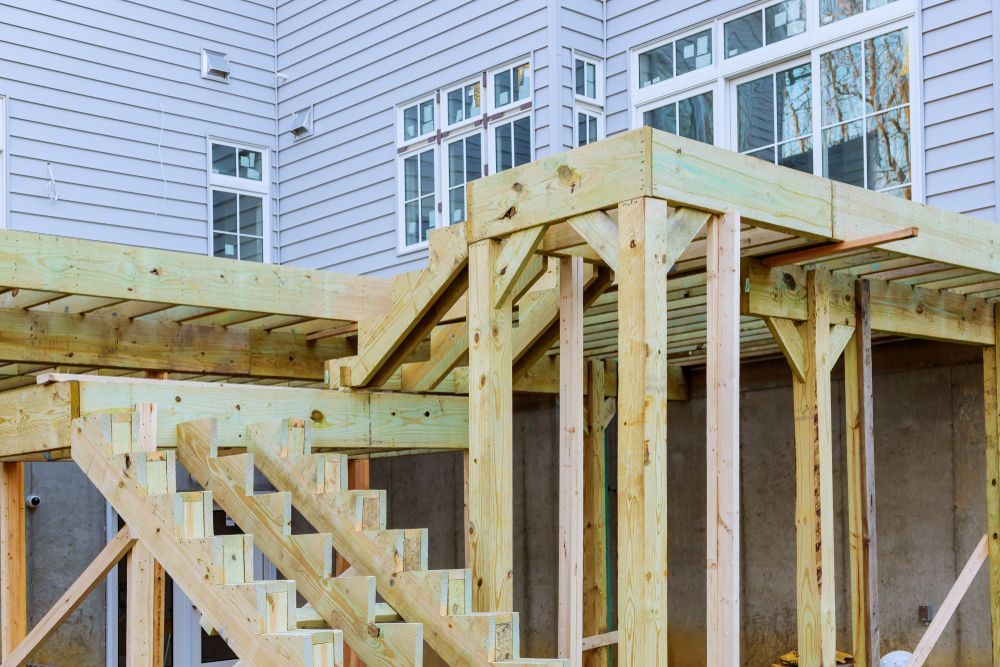 Wooden deck construction with stairs, supports, and partial upper level, next to a light gray house.