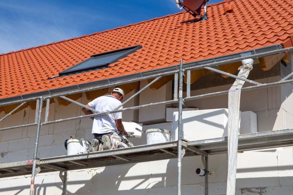 Construction worker on scaffolding, applying material to a white wall beneath a red-tiled roof. Blue sky.