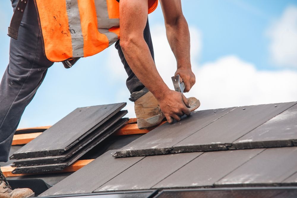 Roofer in orange vest installing dark tiles on a roof under a partly cloudy sky.