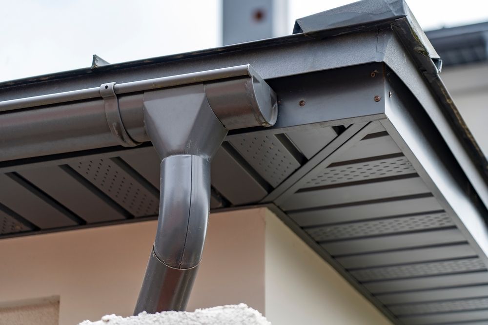 Gray gutter system on a building's roof with downspout, angled corner, and vented soffit paneling.