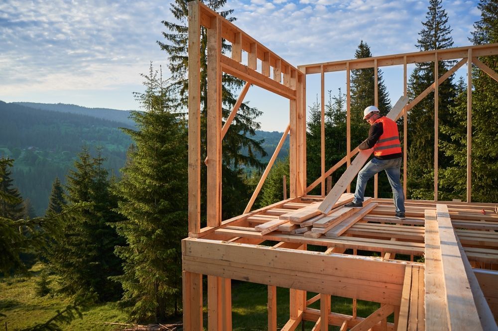Construction worker framing a wooden house, orange vest, mountain backdrop.