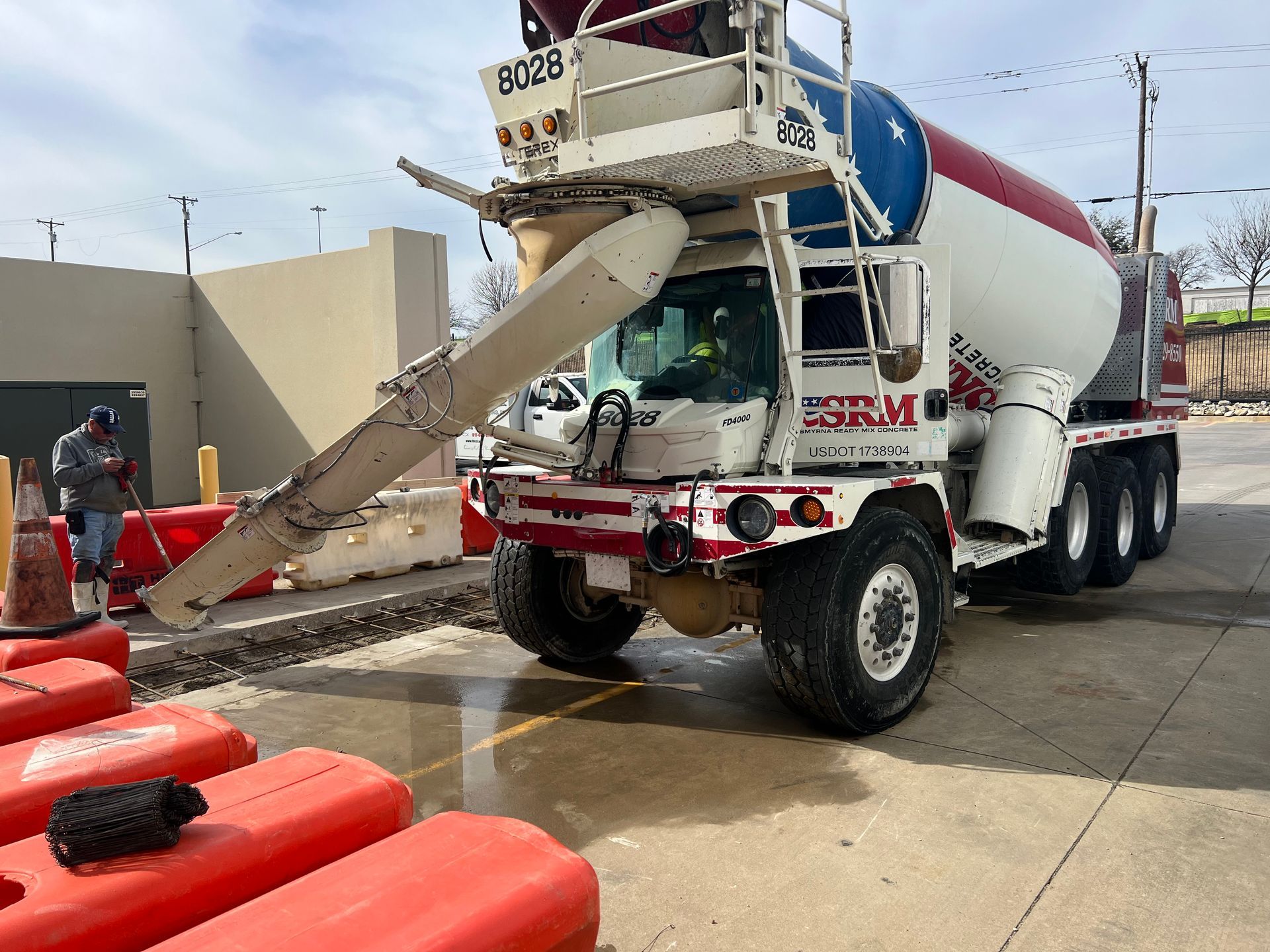 A concrete mixer truck parked on a paved lot with a worker nearby, shielded by orange construction barriers.