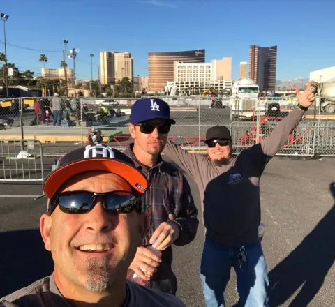 Three people smile for a selfie at an outdoor construction site with the Las Vegas Strip skyline in the background.
