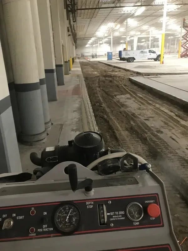 A view from behind the controls of construction machinery, facing a dirt path inside a large, industrial warehouse.