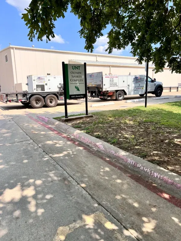 A white UNT service truck and trailer parked in front of a building with a campus sign on a sunny day.