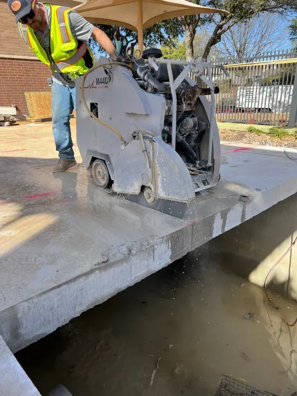 A construction worker operates a walk-behind concrete saw on a slab edge above a muddy excavation.
