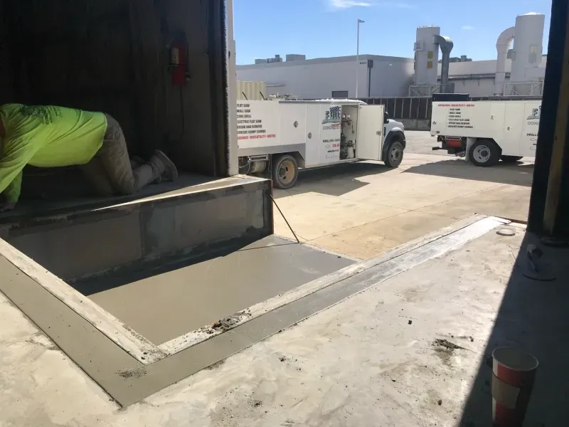 A worker in a high-visibility lime shirt works at a building entrance with freshly poured concrete, two service trucks.