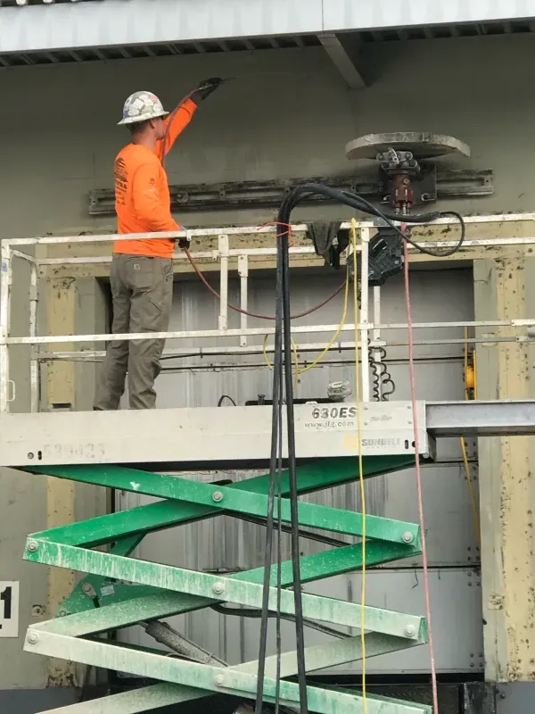 A worker in a bright orange shirt and hard hat stands on a scissor lift, pointing toward a wall section near equipment.