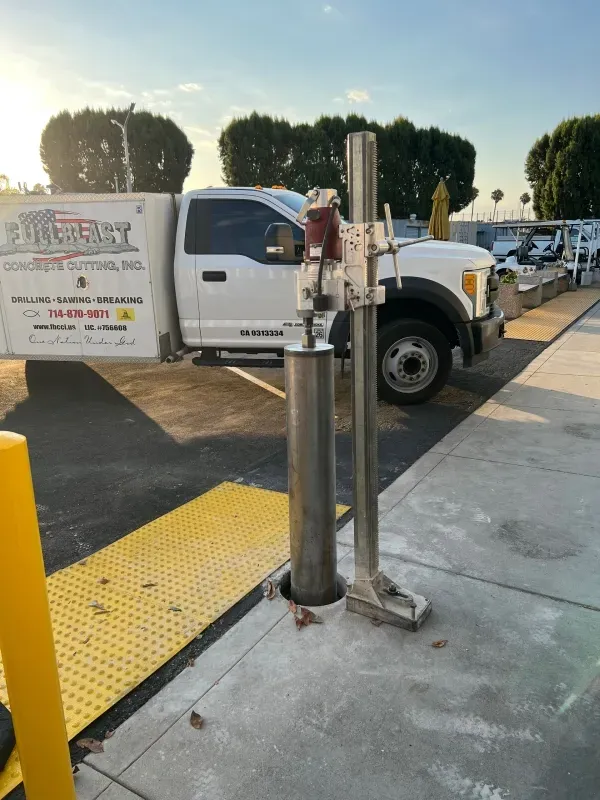 A diamond core drill stand is positioned on a concrete sidewalk, with a service truck parked in the background.