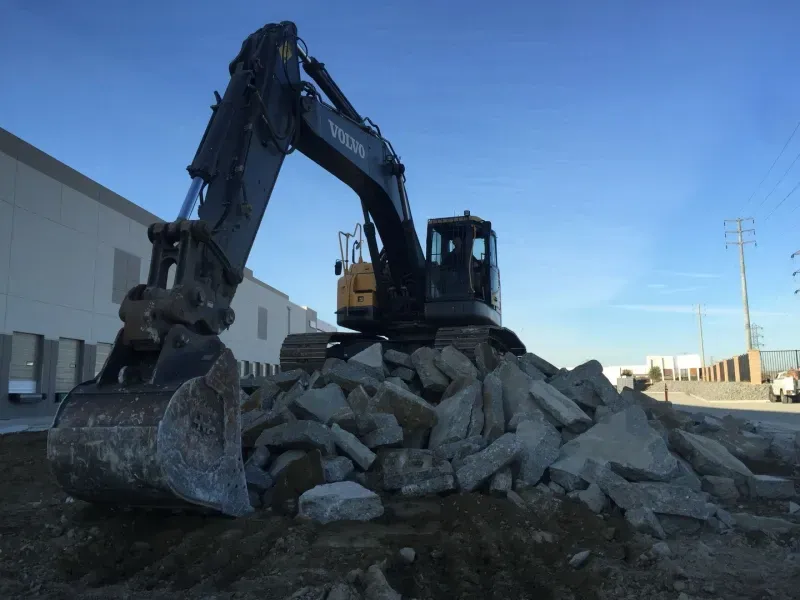 A black excavator sits atop a pile of concrete rubble at a construction site against a clear blue sky.