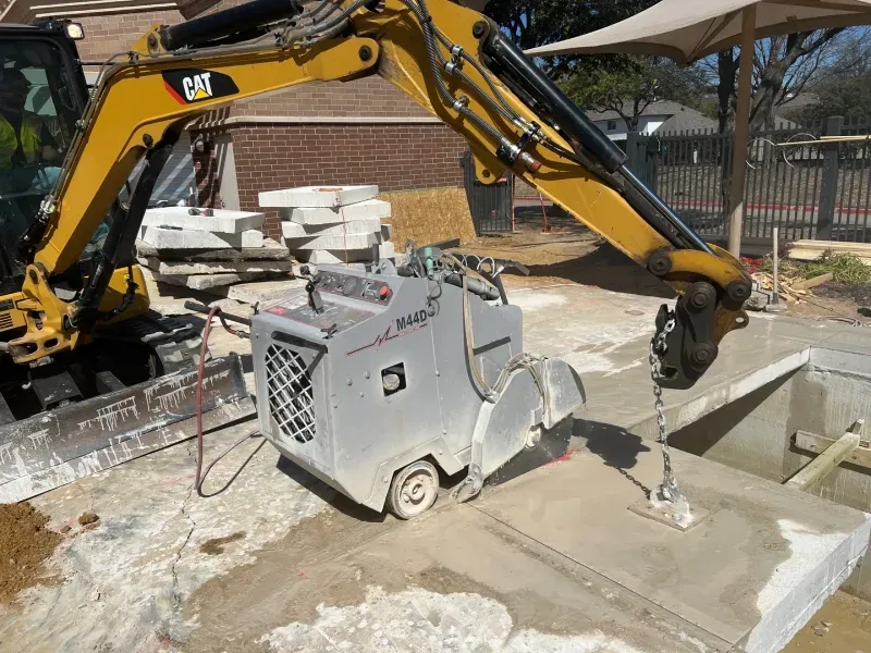 A yellow Caterpillar excavator arm holds a silver walk-behind concrete saw on a construction site.