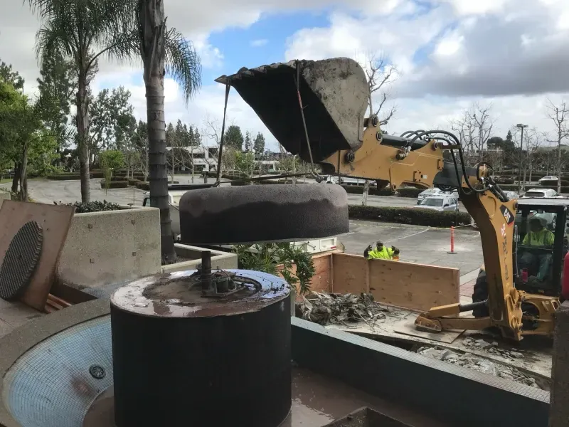A yellow backhoe lifts a circular concrete stone piece over a fountain base at a construction site.