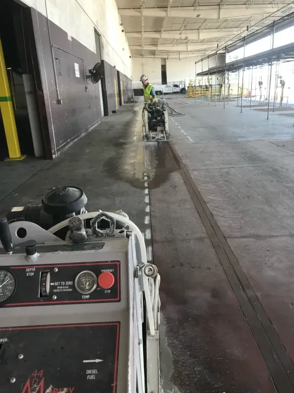 A worker operates a walk-behind floor cleaning machine in a large warehouse, leaving a clean track on the concrete floor.