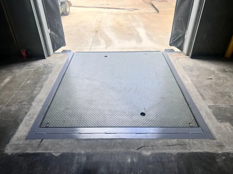 A metal loading dock leveler set into a concrete floor at an open warehouse bay door.