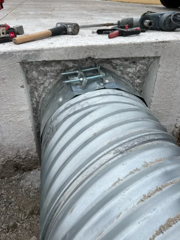A large, corrugated metal pipe secured with a metal clamp and bolts inside a concrete opening at a construction site.