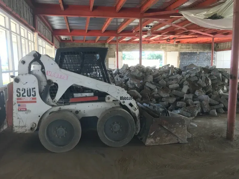 A white Bobcat S205 skid steer loader parked indoors in front of a large pile of rubble and concrete debris.