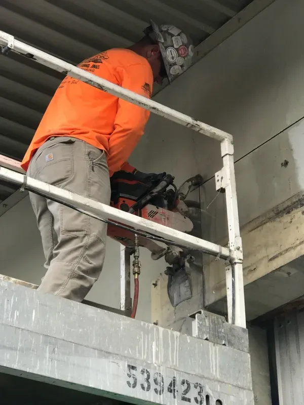 A worker in a high-visibility orange long-sleeve shirt uses a concrete saw while standing on a lift platform.