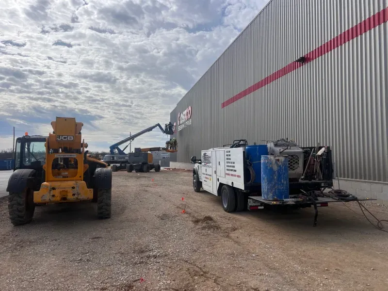 A yellow telehandler, service truck, and boom lift work on the side of a large metal warehouse building under a cloudy sky.