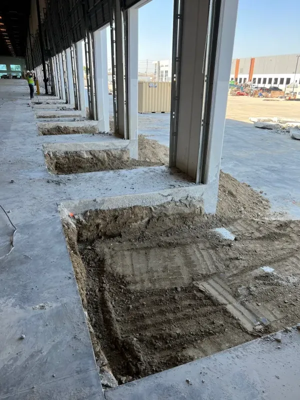 An indoor warehouse site shows rows of concrete column foundations with excavated soil areas along an exterior wall.