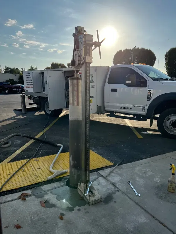 A concrete core drill rig is positioned on a sidewalk next to a white service truck in a parking lot on a sunny day.