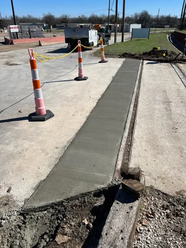 A newly poured concrete sidewalk segment under construction, cordoned off by orange safety cones in a parking lot.
