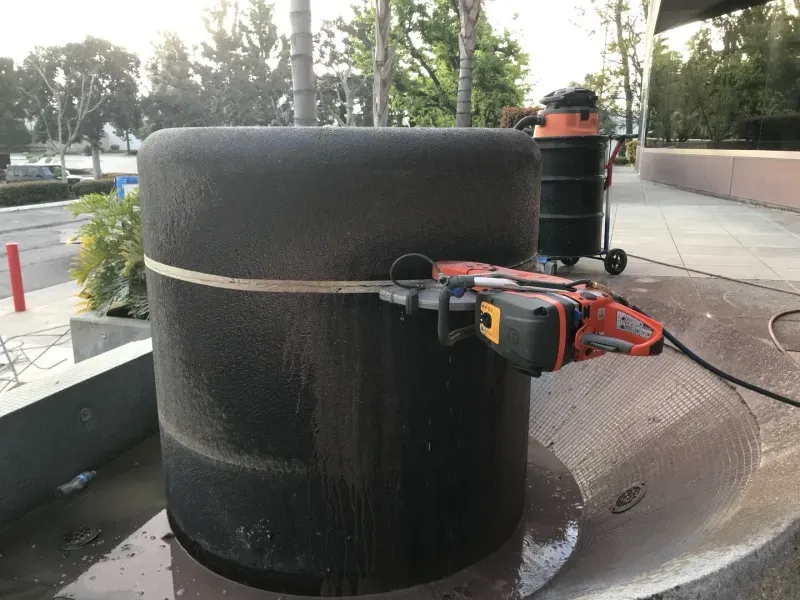 A handheld concrete saw cutting a circular stone planter outdoors with a vacuum cleaner nearby for dust control.