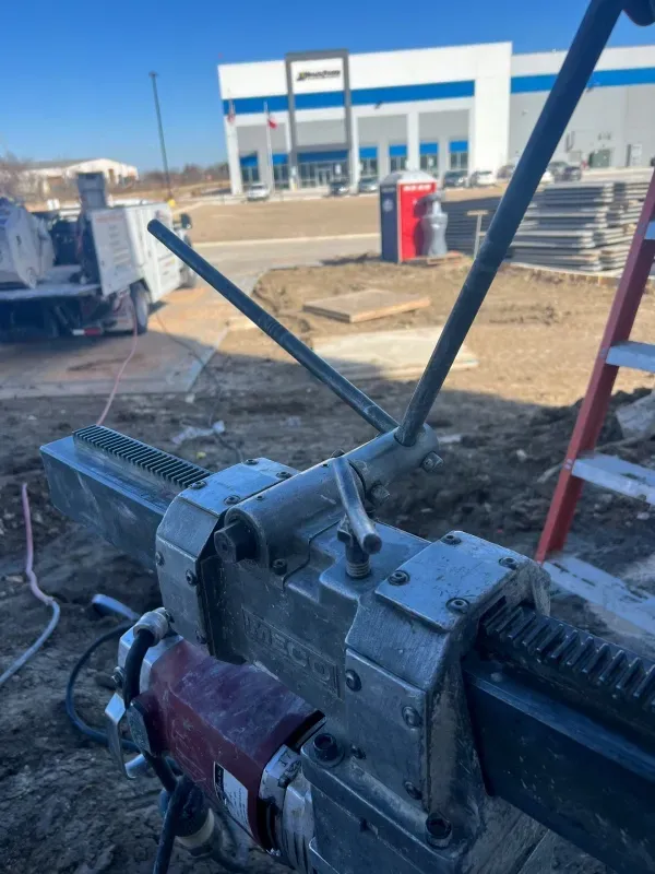 A close-up view of a metal core drilling machine on a construction site with a large building in the background.