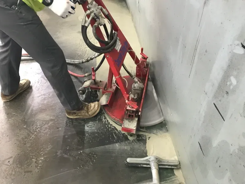 A person uses a red industrial floor saw to cut along the edge of a concrete wall.