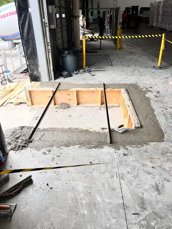 A construction site floor with a rectangular opening framed by wooden boards and freshly poured concrete edges.