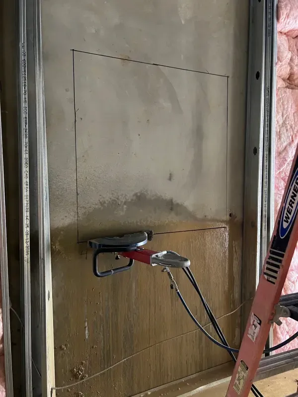 A drywall saw cuts a square hole in a wall section between metal studs, with a red ladder visible on the right.