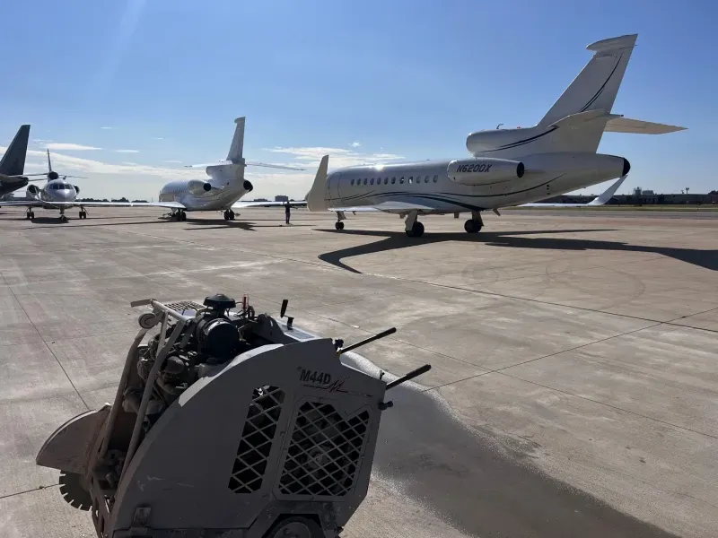 A construction saw sits on an airport tarmac near several parked private jets under a clear blue sky.