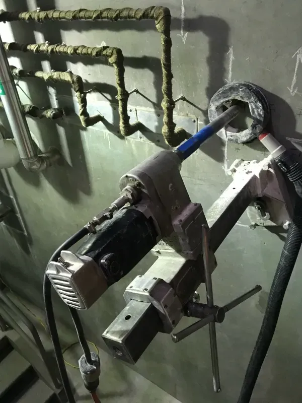 A power tool core drill is mounted on a stand, currently drilling a hole into a gray concrete wall with exposed pipes.