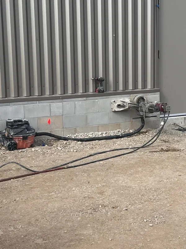 A diamond core drilling rig mounted on a concrete block wall next to a portable vacuum unit on a gravel construction site.