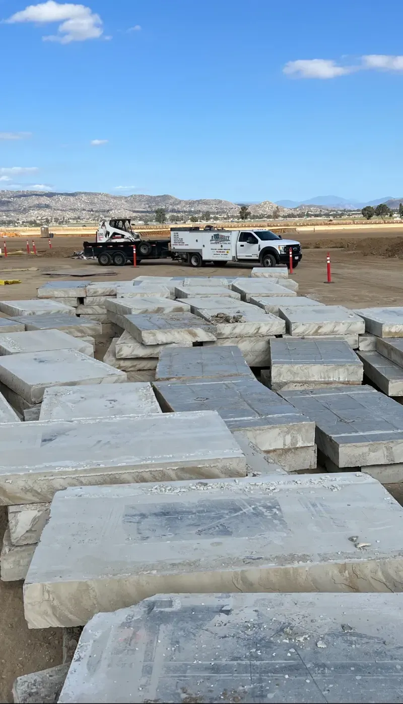 A large pile of concrete slabs sits in a construction field under a blue sky, with a work truck parked in the distance.