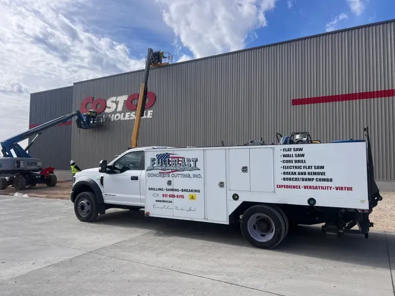 A white work truck parked in front of a Costco building while workers use a lift to install signage on the exterior wall.