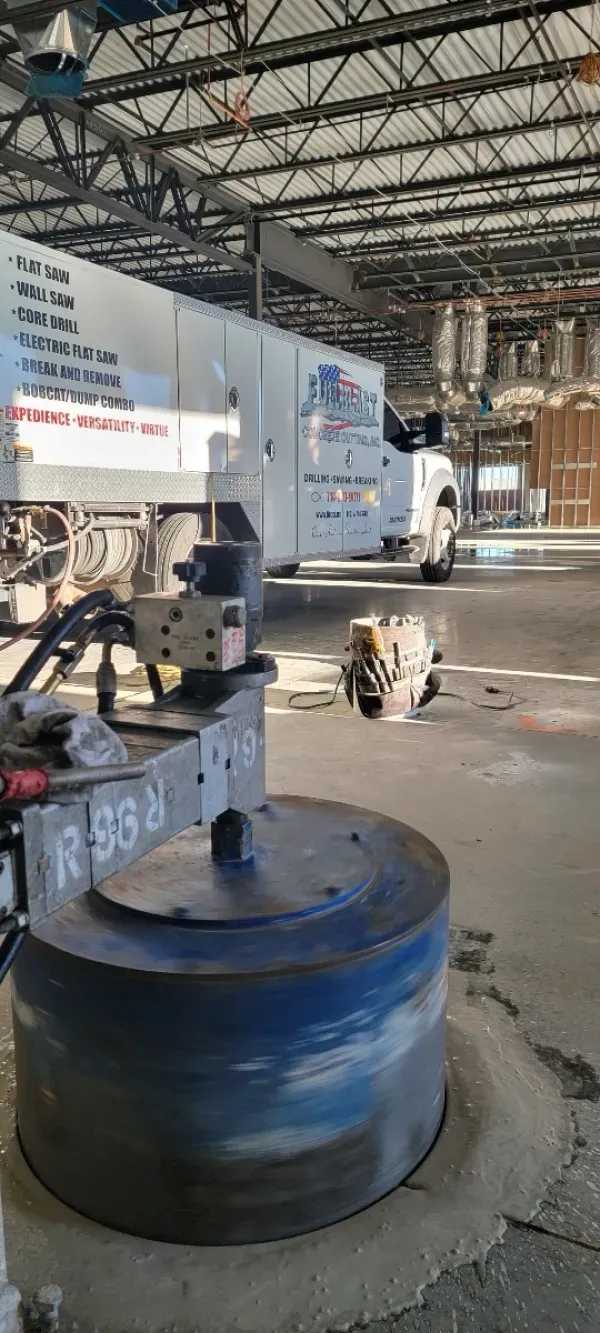 A large circular drill bit sits on a concrete floor in an industrial facility, with a utility truck parked in the back.