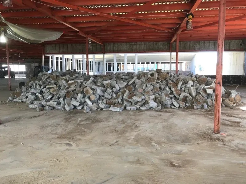 A large pile of broken concrete chunks sits on a dusty floor inside an open-air industrial building.