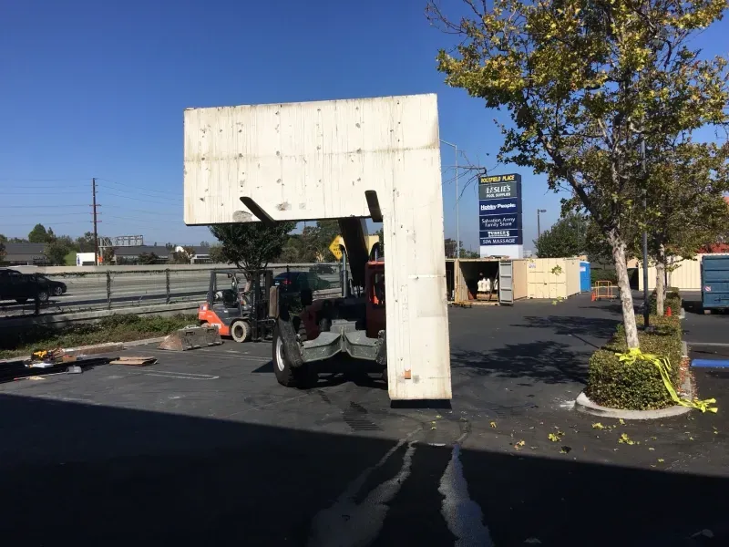 A forklift carries a large, L-shaped white wooden structure across a parking lot on a sunny day.