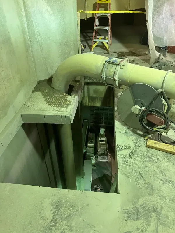 A circular saw sits on a concrete floor next to a rectangular opening containing a vertical pipe and construction debris.