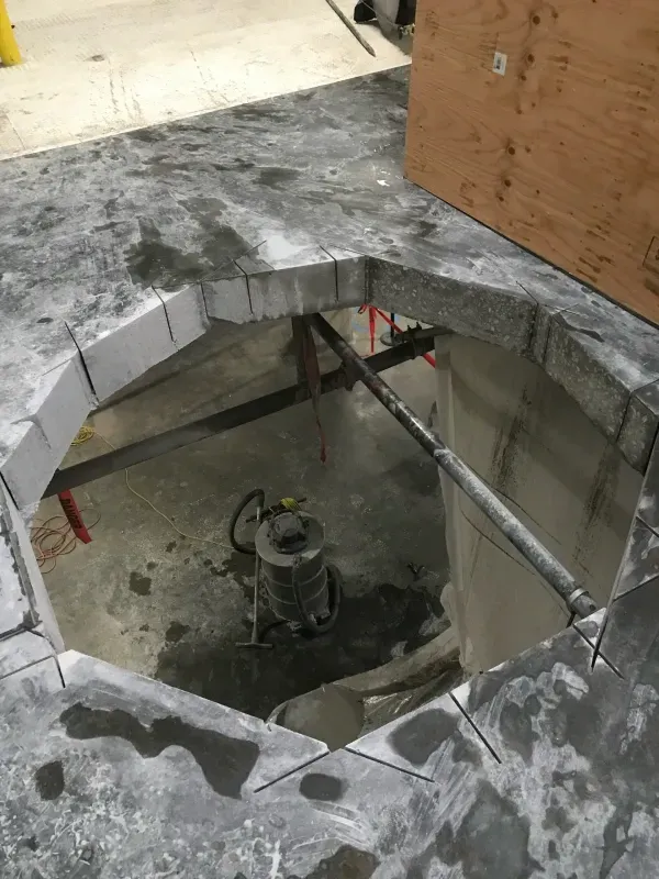 A hole cut into a gray stone slab floor, showing construction equipment and a metal pipe below.
