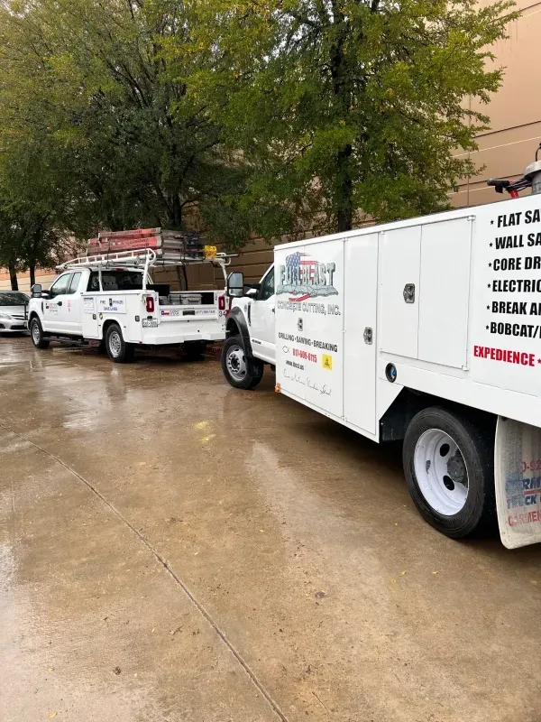 Two white utility trucks parked on a wet concrete surface in front of a tree-lined building.