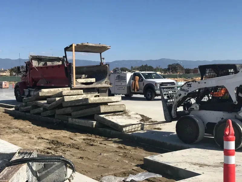 A red dump truck and a white skid steer loader on a construction site, with a pile of concrete slabs in the foreground.