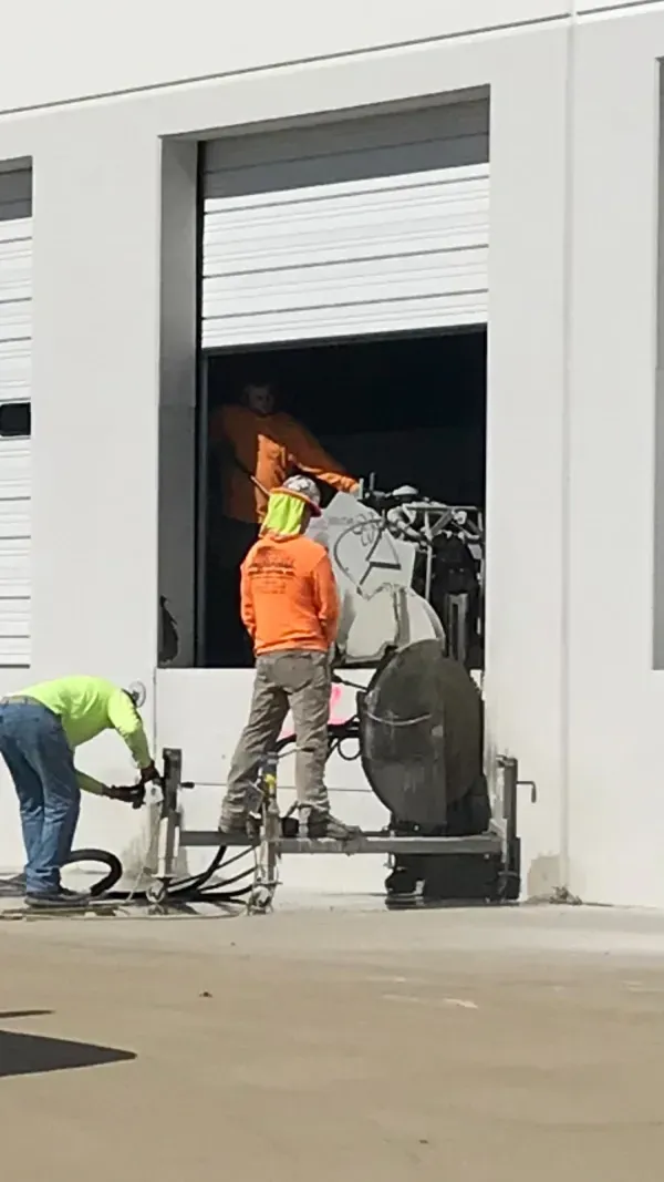 Construction workers in high-visibility orange and lime green shirts operate machinery in a building's open loading bay.